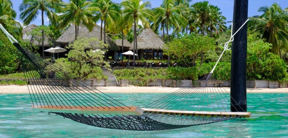 Hammock floating above turquoise water near a sandy beach, surrounded by lush green palm trees and a thatched-roof beachfront resort. Peaceful and tropical.