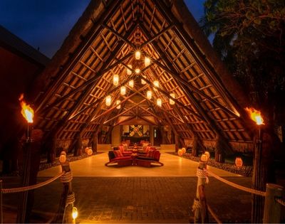 A-frame entryway with thatched roof lit by warm hanging lanterns and torches. Inside, plush red seating creates a cozy, inviting atmosphere.