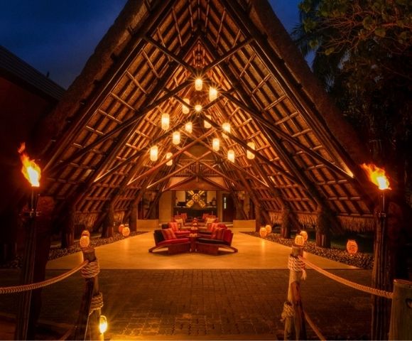 A-frame entryway with thatched roof lit by warm hanging lanterns and torches. Inside, plush red seating creates a cozy, inviting atmosphere.