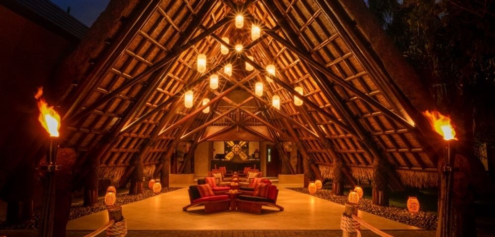 A-frame entryway with thatched roof lit by warm hanging lanterns and torches. Inside, plush red seating creates a cozy, inviting atmosphere.