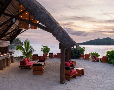 Beachfront lounge with red-cushioned wooden chairs on sandy shore. Thatched roof, sunset sky, ocean view, lush greenery, calm and inviting ambiance.