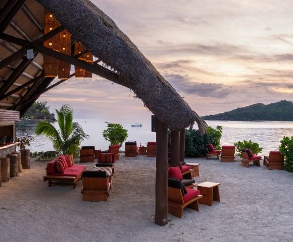Beachfront lounge with red-cushioned wooden chairs on sandy shore. Thatched roof, sunset sky, ocean view, lush greenery, calm and inviting ambiance.