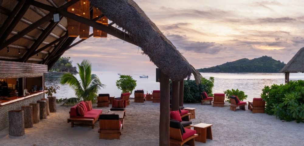 Beachfront lounge with red-cushioned wooden chairs on sandy shore. Thatched roof, sunset sky, ocean view, lush greenery, calm and inviting ambiance.