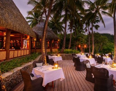Tropical evening setting with a rustic restaurant, candlelit tables on a wooden deck, surrounded by palm trees; a serene and romantic ambiance.