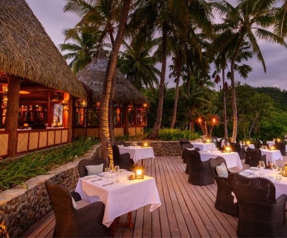 Tropical evening setting with a rustic restaurant, candlelit tables on a wooden deck, surrounded by palm trees; a serene and romantic ambiance.