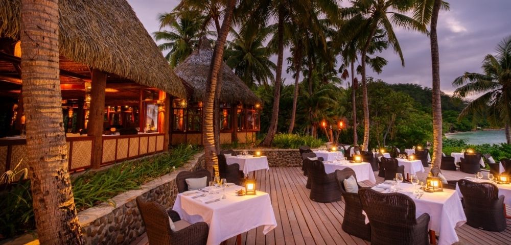 Tropical evening setting with a rustic restaurant, candlelit tables on a wooden deck, surrounded by palm trees; a serene and romantic ambiance.