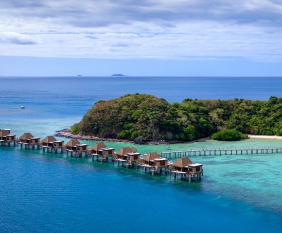 Overwater bungalows stretch along a teal ocean beside a lush, green island. A serene, tropical scene with a clear blue sky and distant islands.