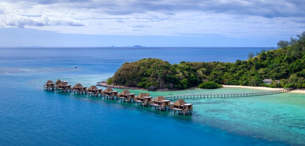 Overwater bungalows stretch along a teal ocean beside a lush, green island. A serene, tropical scene with a clear blue sky and distant islands.
