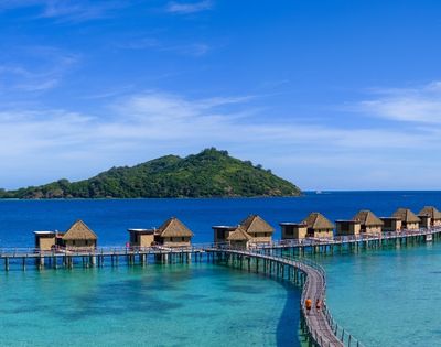 Overwater bungalows with thatched roofs extend along a curved boardwalk above turquoise waters, with a lush green island under a clear blue sky.