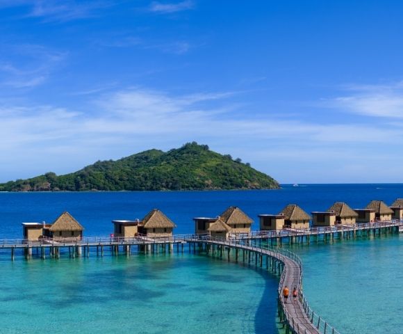 Overwater bungalows with thatched roofs extend along a curved boardwalk above turquoise waters, with a lush green island under a clear blue sky.