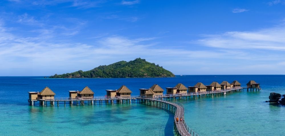 Overwater bungalows with thatched roofs extend along a curved boardwalk above turquoise waters, with a lush green island under a clear blue sky.