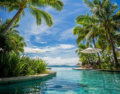 Tropical paradise scene with a clear pool surrounded by lush palm trees. Two white umbrellas provide shade, overlooking an ocean view under a bright blue sky.