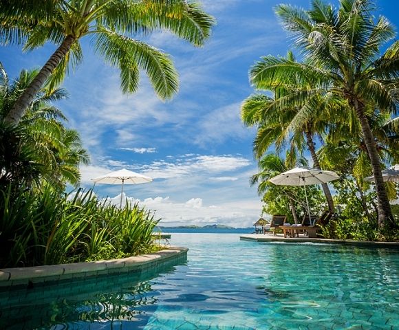 Tropical paradise scene with a clear pool surrounded by lush palm trees. Two white umbrellas provide shade, overlooking an ocean view under a bright blue sky.