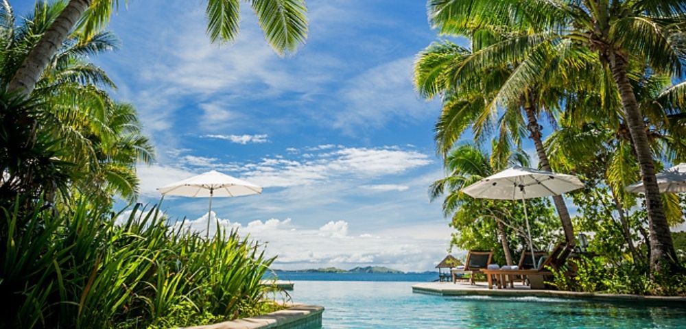 Tropical paradise scene with a clear pool surrounded by lush palm trees. Two white umbrellas provide shade, overlooking an ocean view under a bright blue sky.