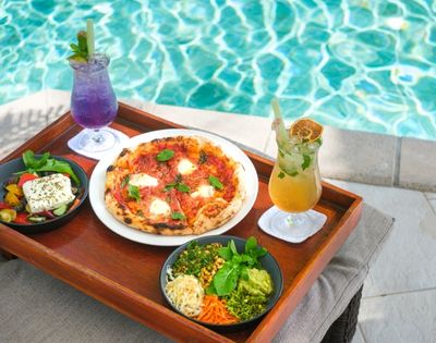 Tray by poolside with fresh pizza, salad, and bowls of colorful vegetables paired with vibrant purple and yellow drinks, evoking a tropical, relaxed vibe.
