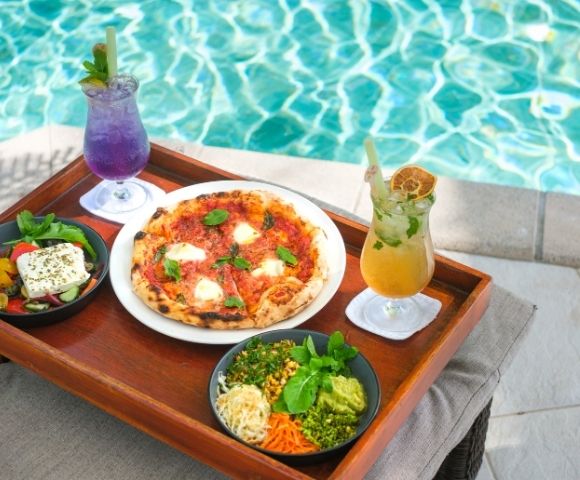 Tray by poolside with fresh pizza, salad, and bowls of colorful vegetables paired with vibrant purple and yellow drinks, evoking a tropical, relaxed vibe.