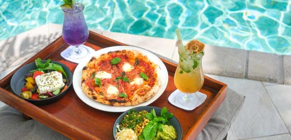 Tray by poolside with fresh pizza, salad, and bowls of colorful vegetables paired with vibrant purple and yellow drinks, evoking a tropical, relaxed vibe.