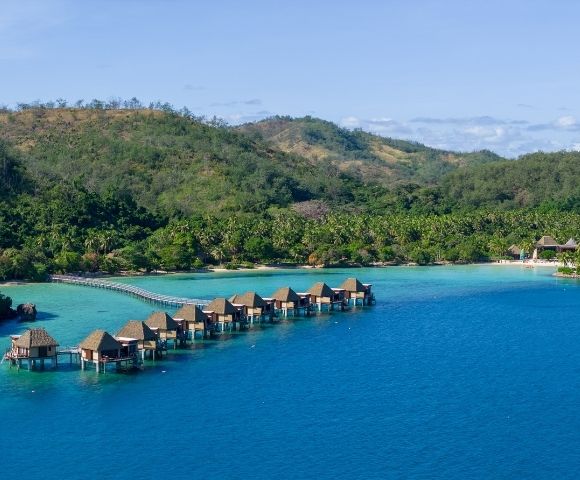 Aerial view of overwater bungalows leading to lush green hills on a tropical island. Clear blue waters and a calm atmosphere evoke tranquility and luxury.