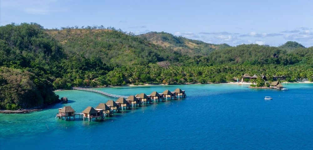 Aerial view of overwater bungalows leading to lush green hills on a tropical island. Clear blue waters and a calm atmosphere evoke tranquility and luxury.
