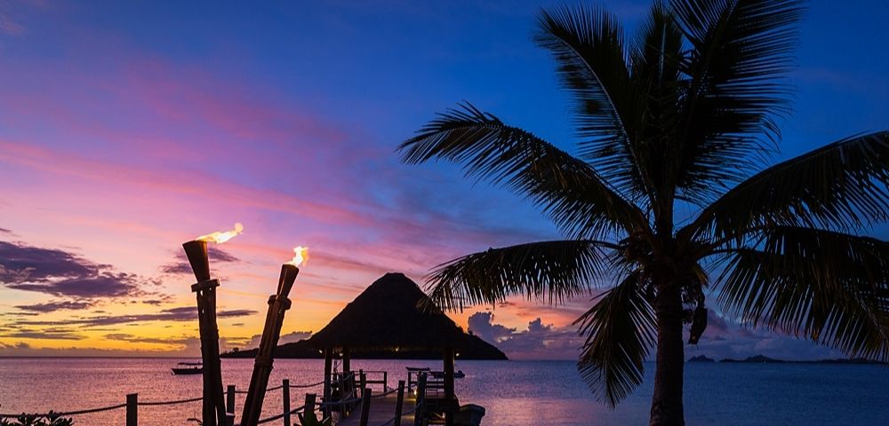 A serene tropical sunset over the ocean with vibrant pink and blue skies. A silhouette of a palm tree and two lit tiki torches enhances the peaceful scene.