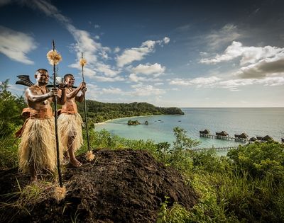Two people in traditional attire stand on a hill overlooking a serene coastal landscape with lush greenery, stilted huts over turquoise waters, and a bright sky.