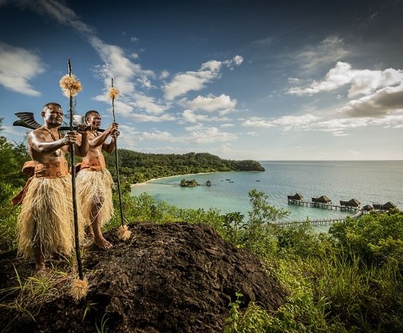 Two people in traditional attire stand on a hill overlooking a serene coastal landscape with lush greenery, stilted huts over turquoise waters, and a bright sky.