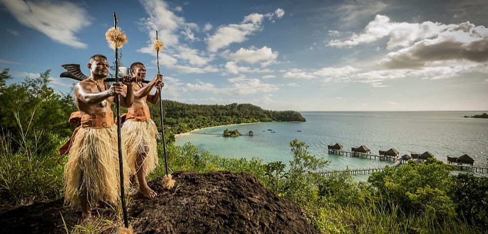 Two people in traditional attire stand on a hill overlooking a serene coastal landscape with lush greenery, stilted huts over turquoise waters, and a bright sky.