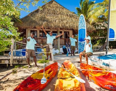 Four smiling men in matching blue shirts stand by colorful kayaks and paddleboards on a sunny beach. Behind them, a thatched-roof hut and palm trees create a tropical vibe.
