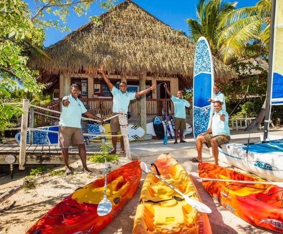Four smiling men in matching blue shirts stand by colorful kayaks and paddleboards on a sunny beach. Behind them, a thatched-roof hut and palm trees create a tropical vibe.