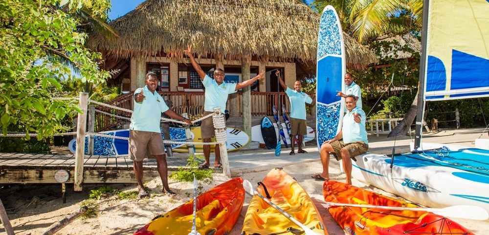 Four smiling men in matching blue shirts stand by colorful kayaks and paddleboards on a sunny beach. Behind them, a thatched-roof hut and palm trees create a tropical vibe.