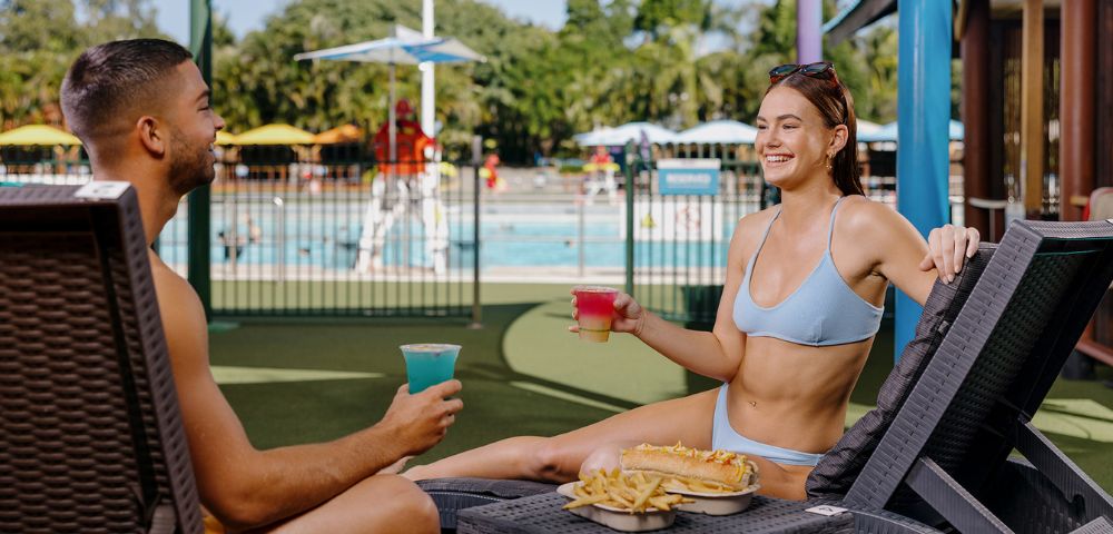 A smiling man and woman in swimwear lounge by a pool, holding colorful drinks. A tray with fries and a hot dog is on a table between them. Sunny, relaxed vibe.