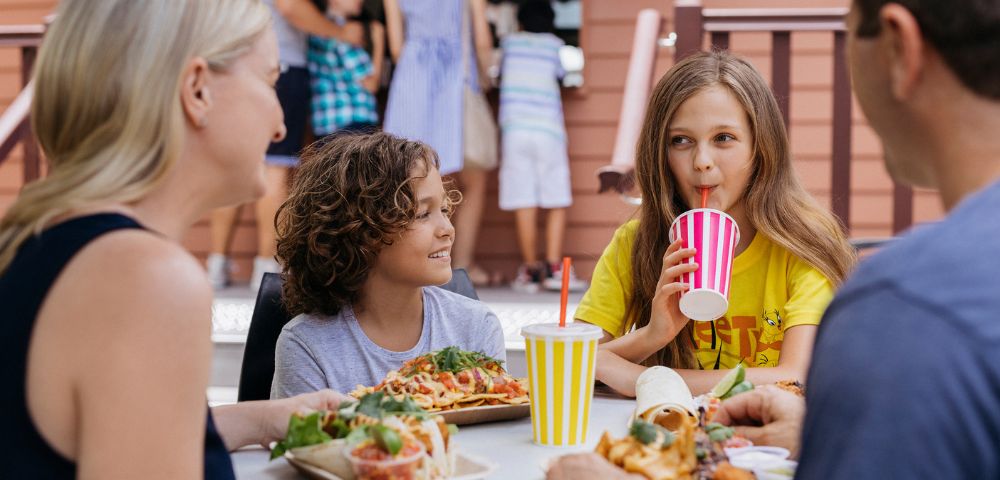 A family enjoys a meal outdoors. A girl in a yellow shirt sips a drink while a boy smiles. Plates of food and drinks are on the table, creating a warm, joyful atmosphere.