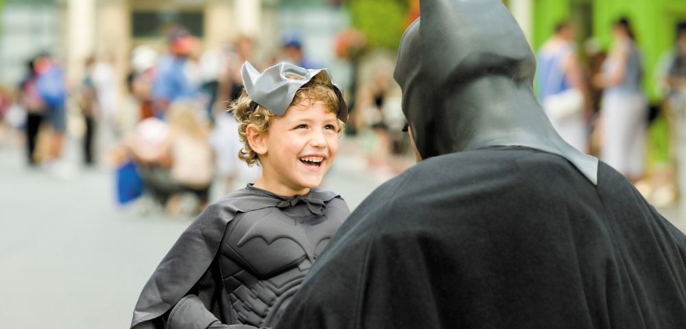A young boy dressed as Batman smiles joyfully at an adult in a Batman costume. They are in a crowded outdoor area, conveying excitement and fun.