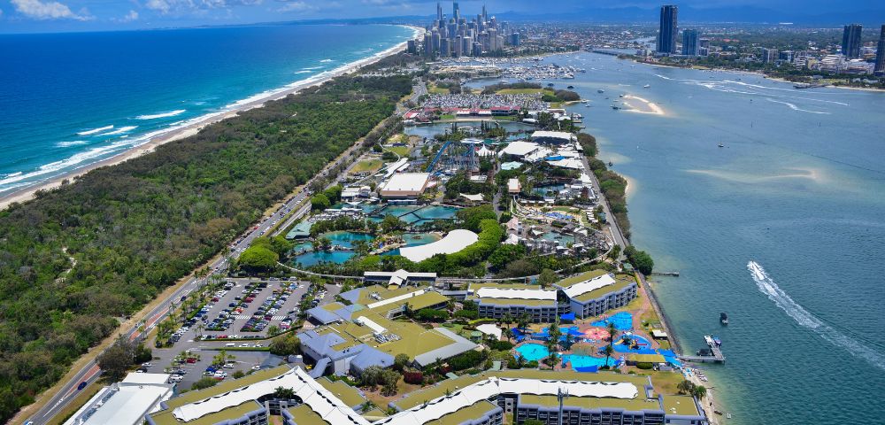 Aerial view of a coastal cityscape with high-rise buildings, lush green parks, and a marina beside a beach. Vibrant blue ocean under a bright sky.