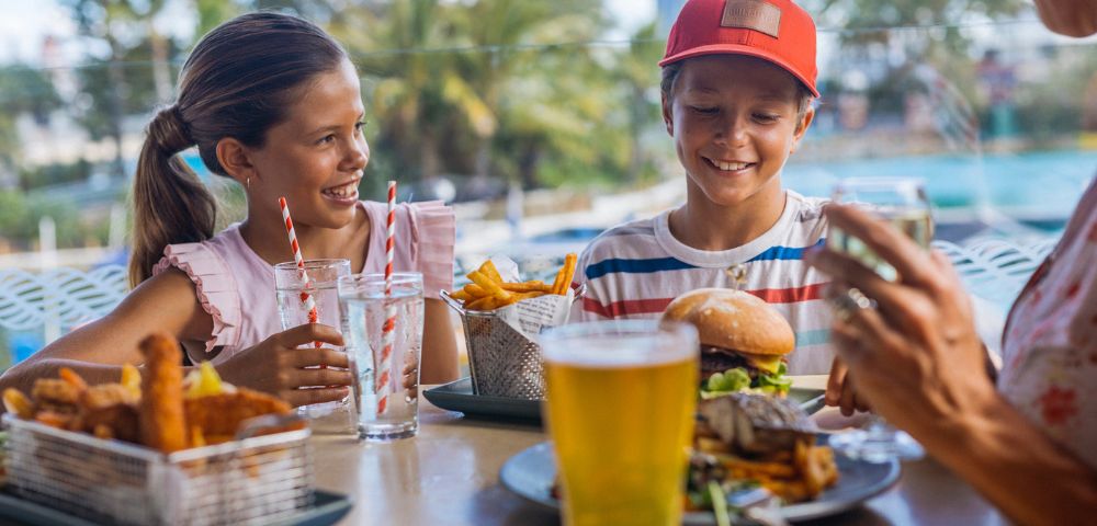 Two children enjoying burgers and fries at an outdoor restaurant table, smiling under sunny skies. Tropical setting. Drinks and vibrant atmosphere.