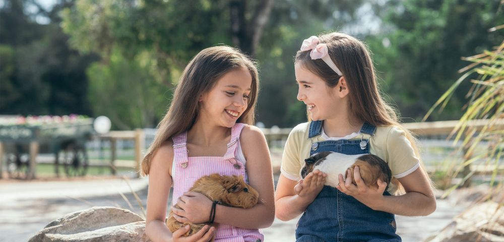 Two smiling girls sit outdoors on rocks, each holding a guinea pig. They wear colorful overalls, have long hair, and are surrounded by greenery, conveying joy.
