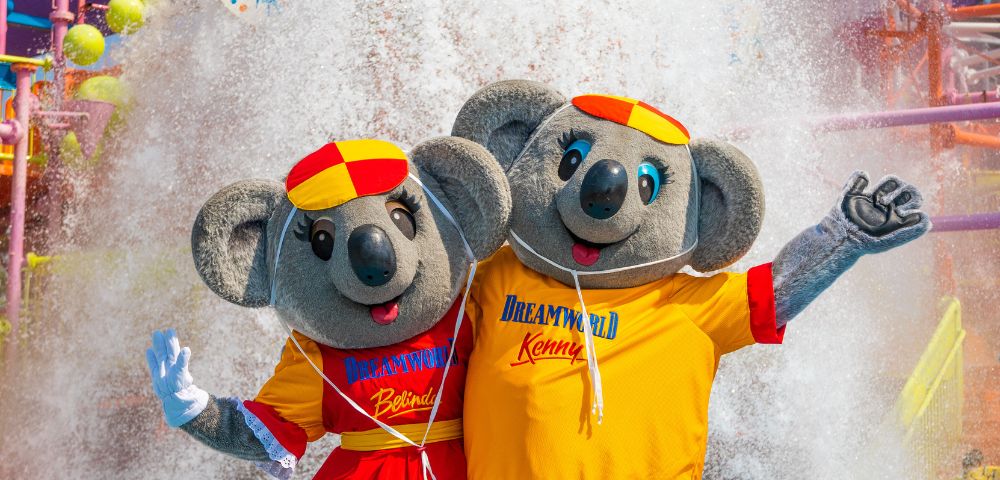 Two cheerful koala mascots in colorful outfits stand joyfully in front of a water spray at an amusement park. They wave with smiles, conveying fun and excitement.