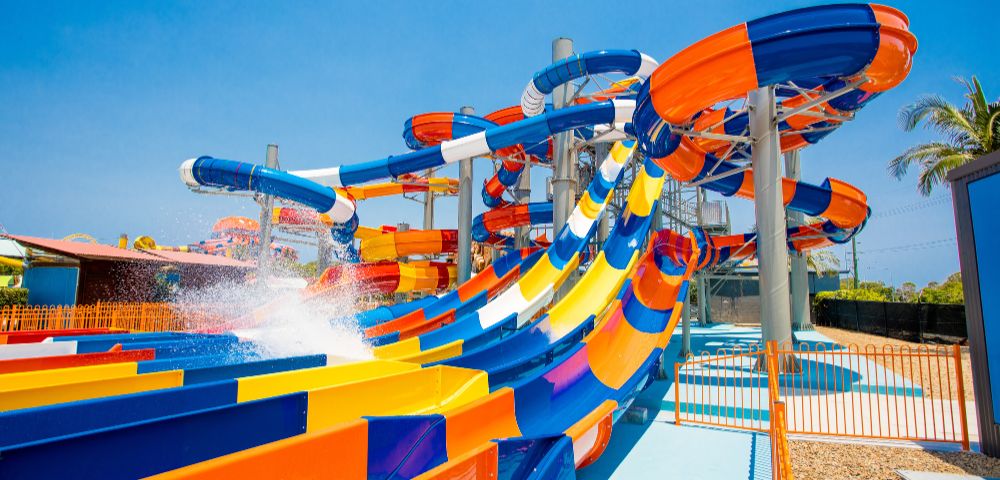 Thrilling water park scene with colorful, intertwined slides in blue, orange, and yellow under a clear blue sky, evoking fun and excitement.