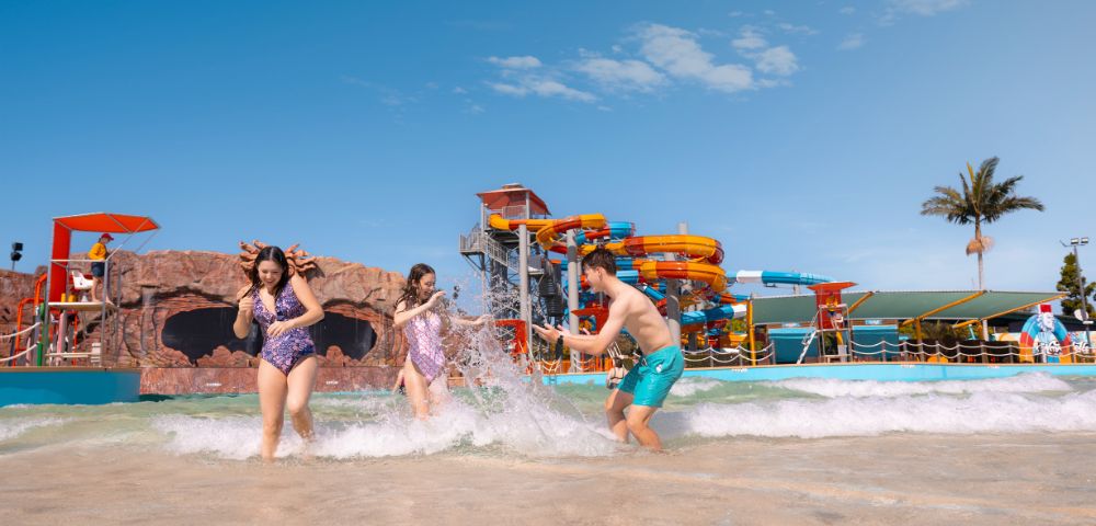 Three people play joyfully in shallow water with splashes around them. Behind, vibrant water slides and palm trees under a clear blue sky create a fun atmosphere.