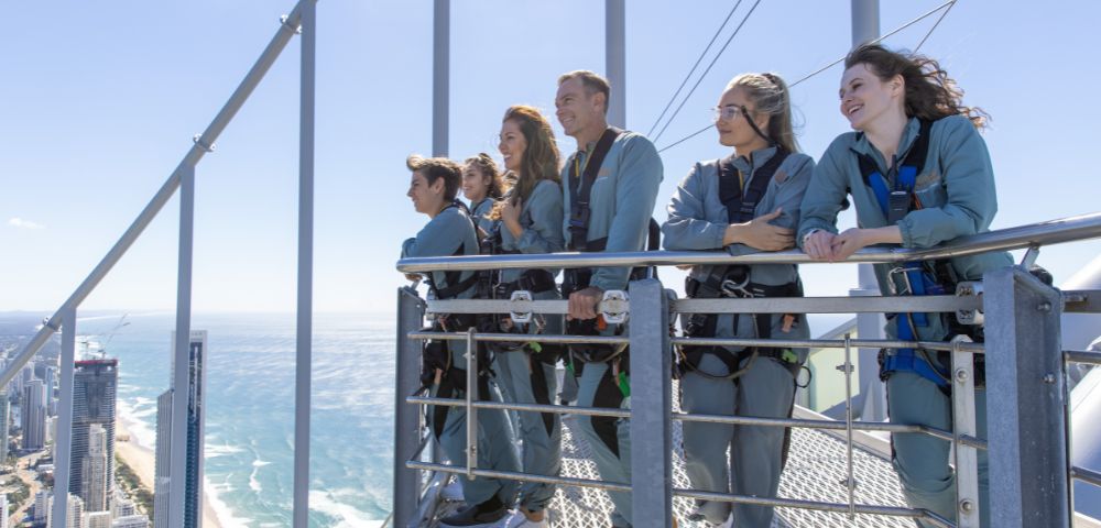 A group of six people, wearing harnesses and blue jumpsuits, stand on a high platform, enjoying a scenic ocean view under a clear blue sky, looking excited.