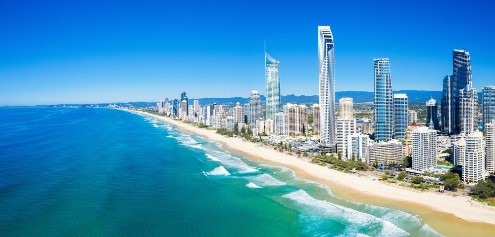 Aerial view of a coastal cityscape, featuring tall skyscrapers lining a sandy beach. The ocean is vibrant blue with gentle waves under a clear sky.