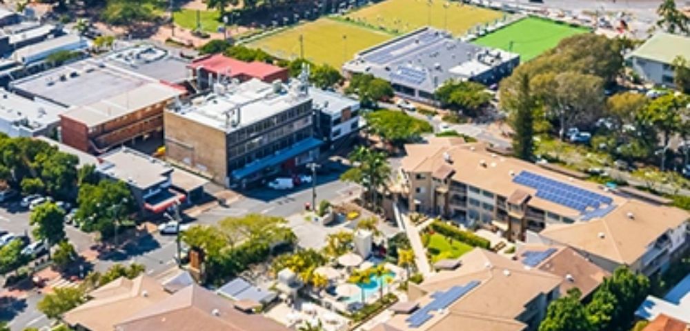 Aerial view of a suburban area with buildings and parking lots surrounded by trees. A vibrant sports field is visible, providing a lively, active atmosphere.