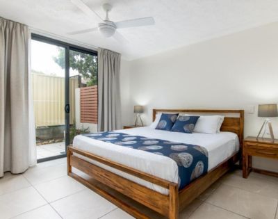 A cozy bedroom with a wooden bed frame, blue and white bedding, flanked by two wooden nightstands and lamps. A sliding glass door leads to a patio.