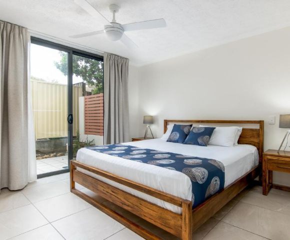A cozy bedroom with a wooden bed frame, blue and white bedding, flanked by two wooden nightstands and lamps. A sliding glass door leads to a patio.