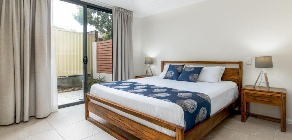 A cozy bedroom with a wooden bed frame, blue and white bedding, flanked by two wooden nightstands and lamps. A sliding glass door leads to a patio.