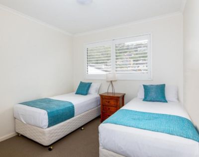 A bright bedroom with two single beds, each with turquoise throws and pillows. A wooden nightstand with a lamp is between them. White walls and a window create an airy feel.