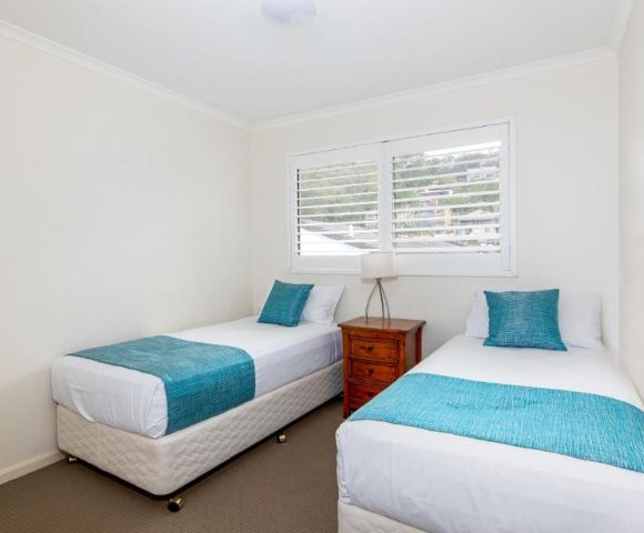 A bright bedroom with two single beds, each with turquoise throws and pillows. A wooden nightstand with a lamp is between them. White walls and a window create an airy feel.