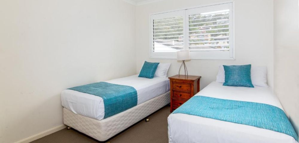A bright bedroom with two single beds, each with turquoise throws and pillows. A wooden nightstand with a lamp is between them. White walls and a window create an airy feel.