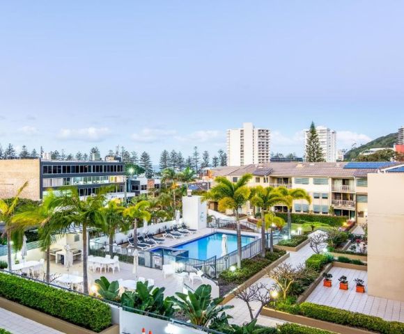 Rooftop view of a modern apartment complex featuring a central swimming pool, surrounded by palm trees and lush greenery. Tall buildings in the background.