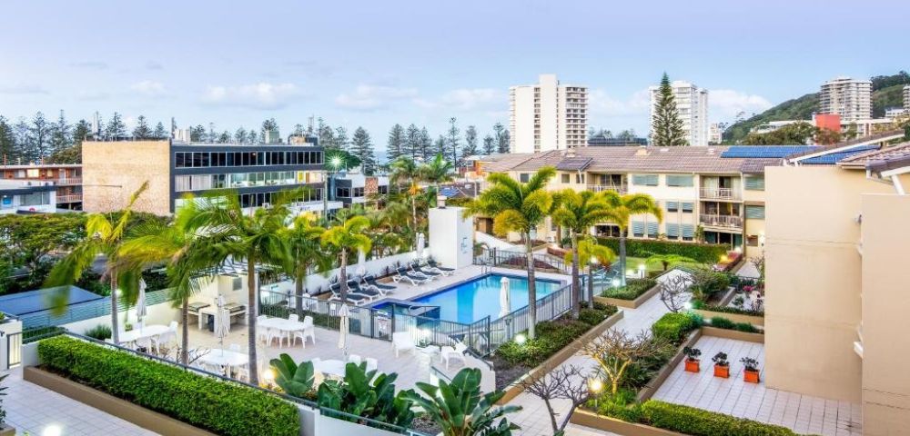 Rooftop view of a modern apartment complex featuring a central swimming pool, surrounded by palm trees and lush greenery. Tall buildings in the background.